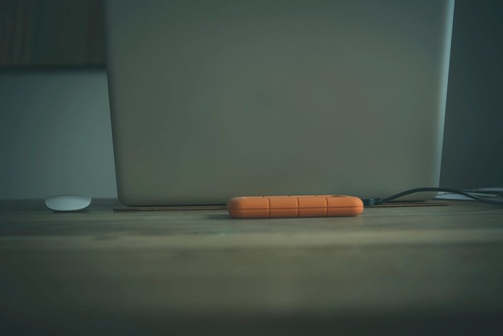 Externe Festplatte in orangefarbener Schutzhülle liegt vor einem aufgeklappten Laptop auf einem Holztisch